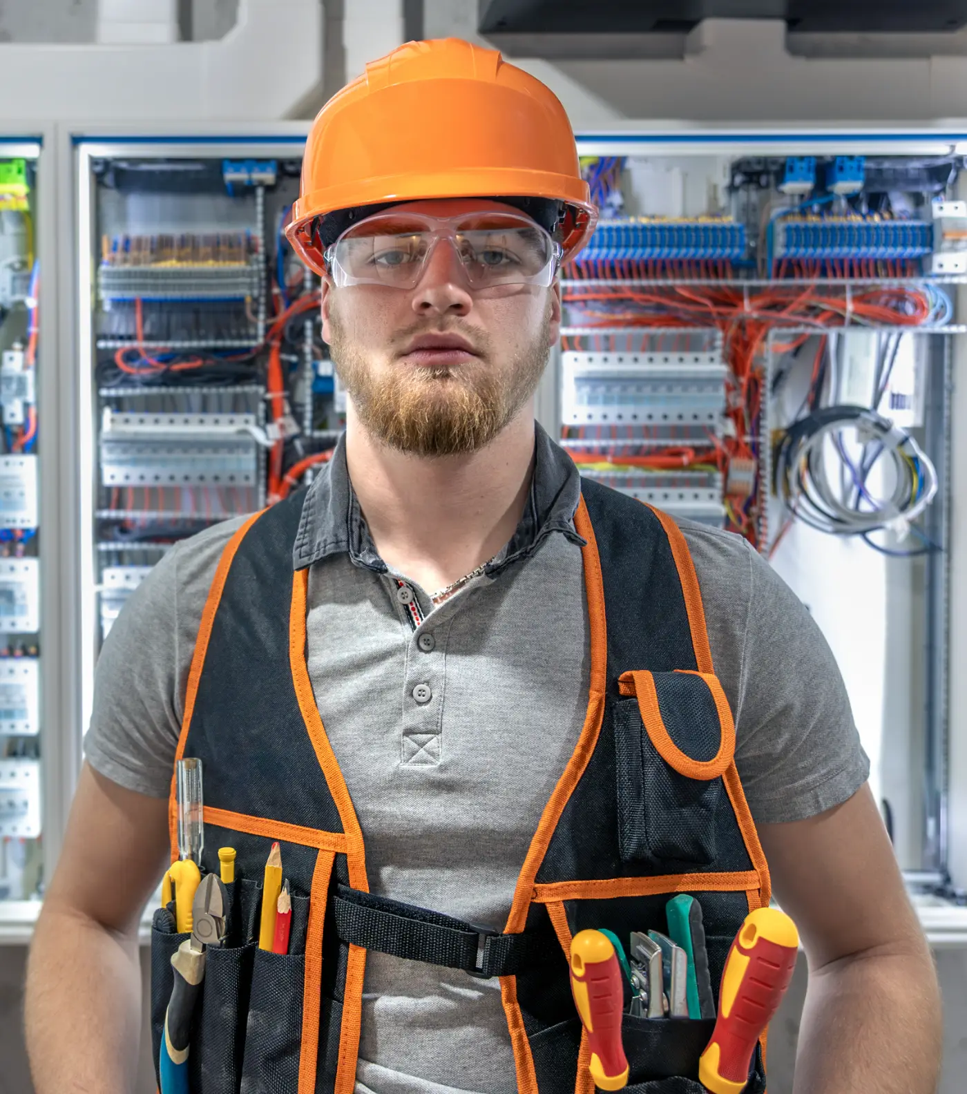 An electrician in a hard hat, safety glasses and a tool vest, standing in front of two open electrical distribution panels.
