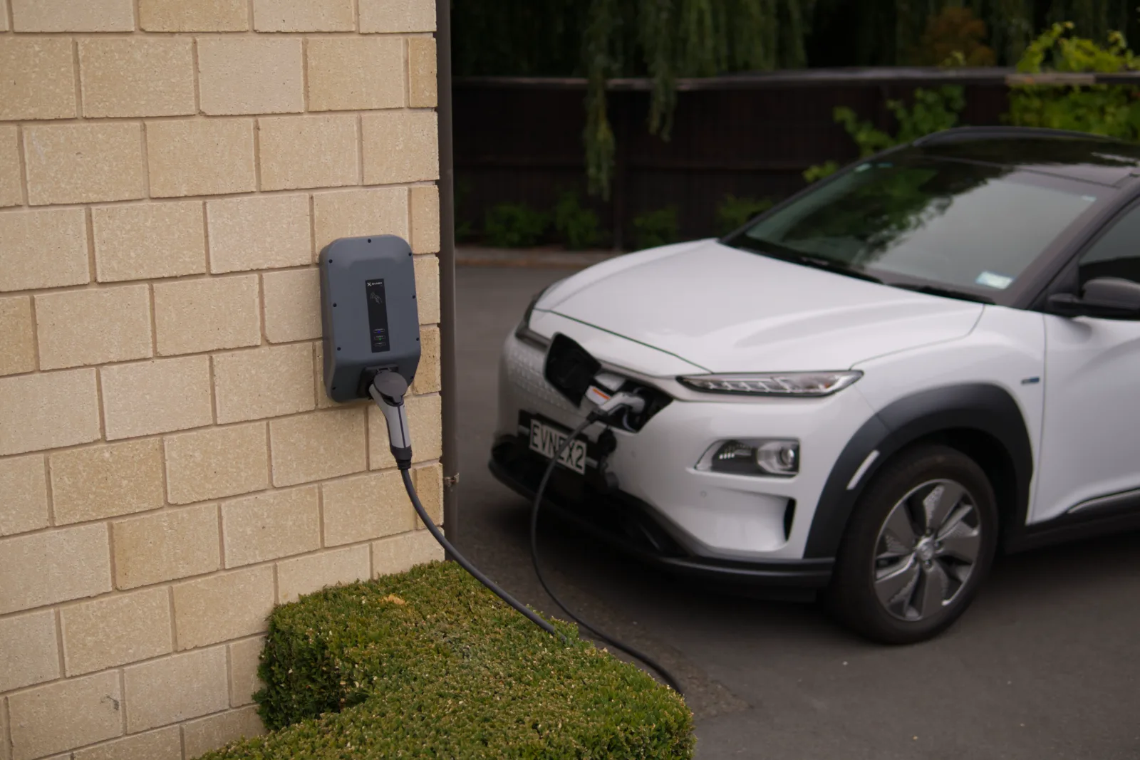 A wall-mounted electric vehicle charge point on the side of a house, its cable plugged into a white car parked on the drive.