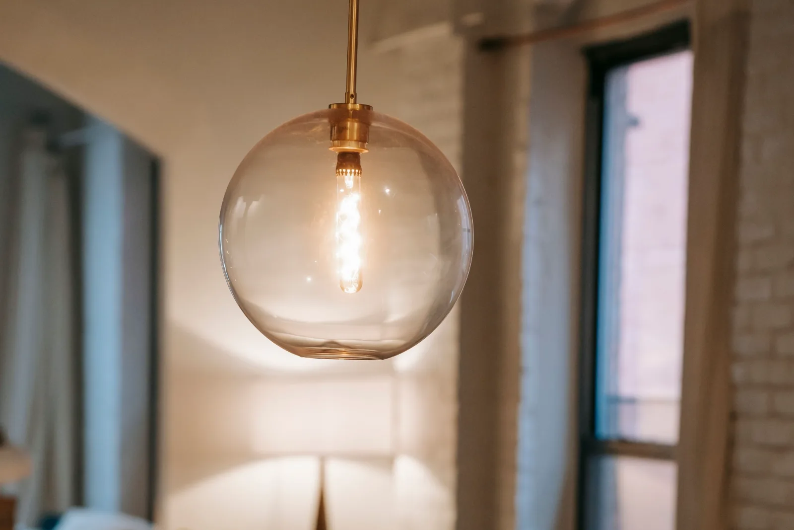 A brass and clear glass globe pendant light hanging on a rod from the ceiling, lit, in a warm living room.